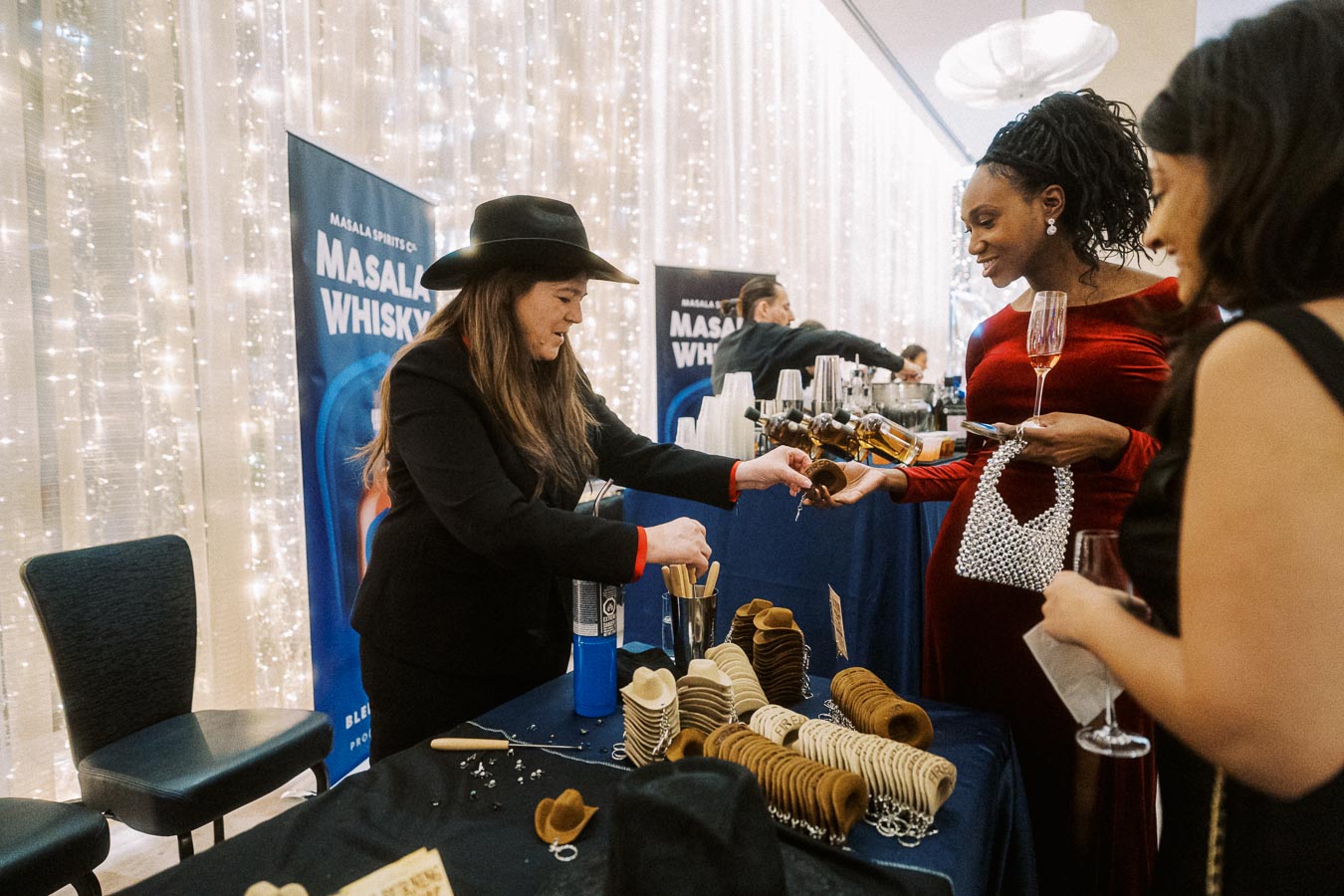 Woman in a black hat serving whiskey samples at an elegant event, with Masala Whiskey signage in the background, as attendees in formal attire enjoy drinks and conversation.