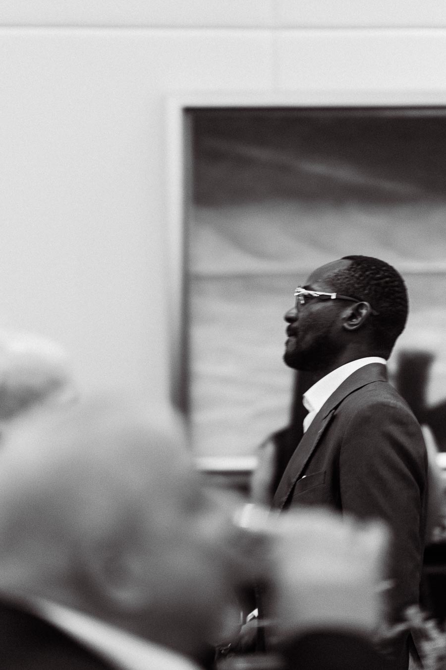 Black and white image of a well-dressed man standing in profile, wearing glasses and a suit jacket, with an art gallery setting in the background.