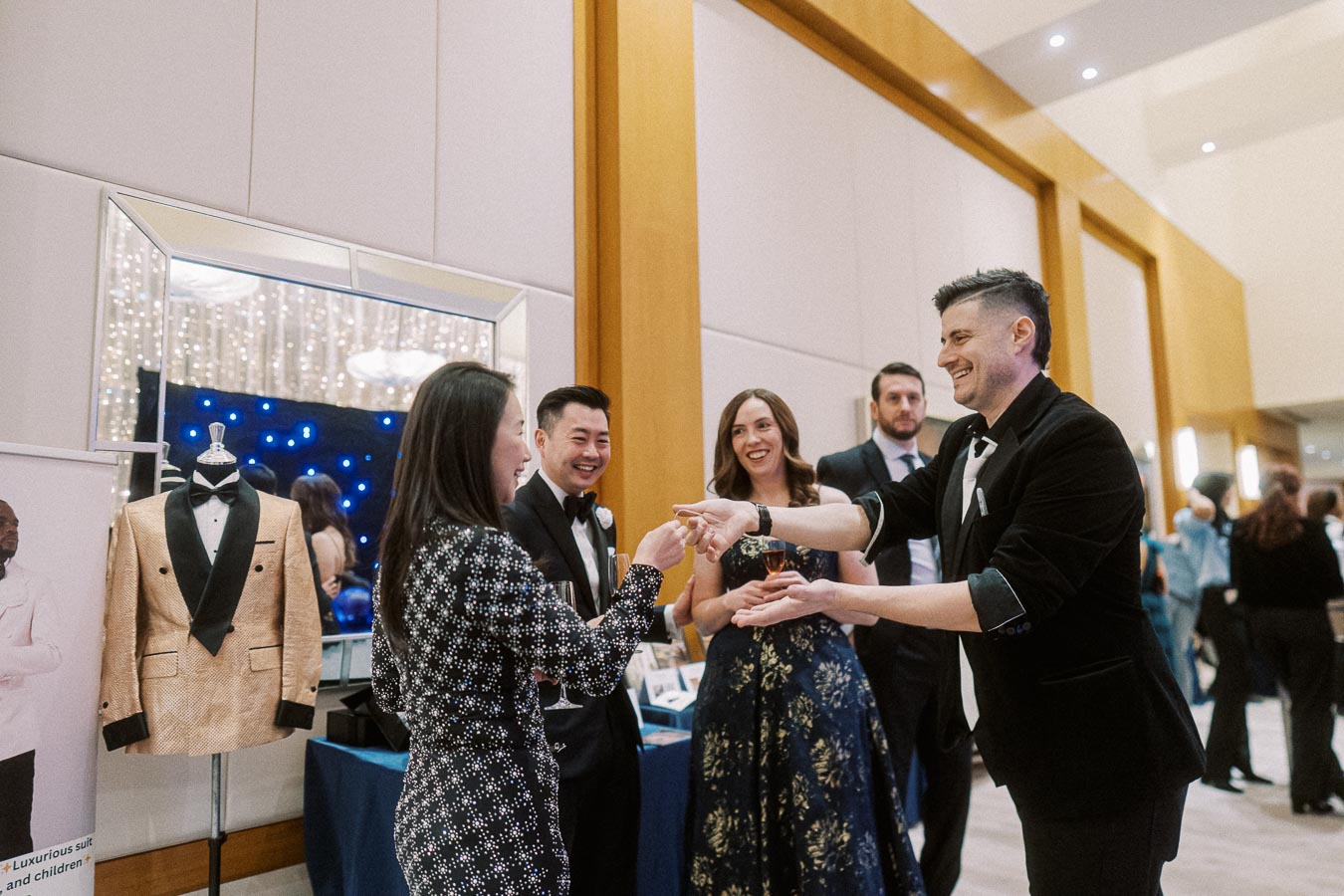 Group of elegantly dressed people enjoying a social event, showcasing a lively interaction and networking atmosphere in a formal setting with a gold tuxedo on display in the background.
