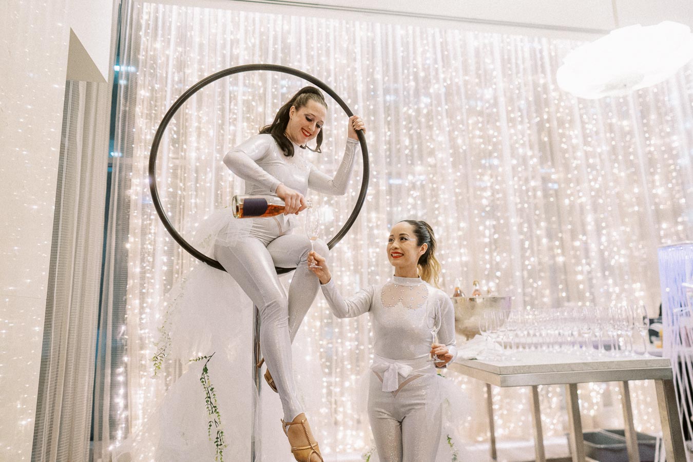 Two performers in shimmering metallic outfits, one sitting on an aerial hoop and pouring champagne for the other, amid a dazzling backdrop of twinkling lights, creating an enchanting and elegant atmosphere at a sophisticated event.