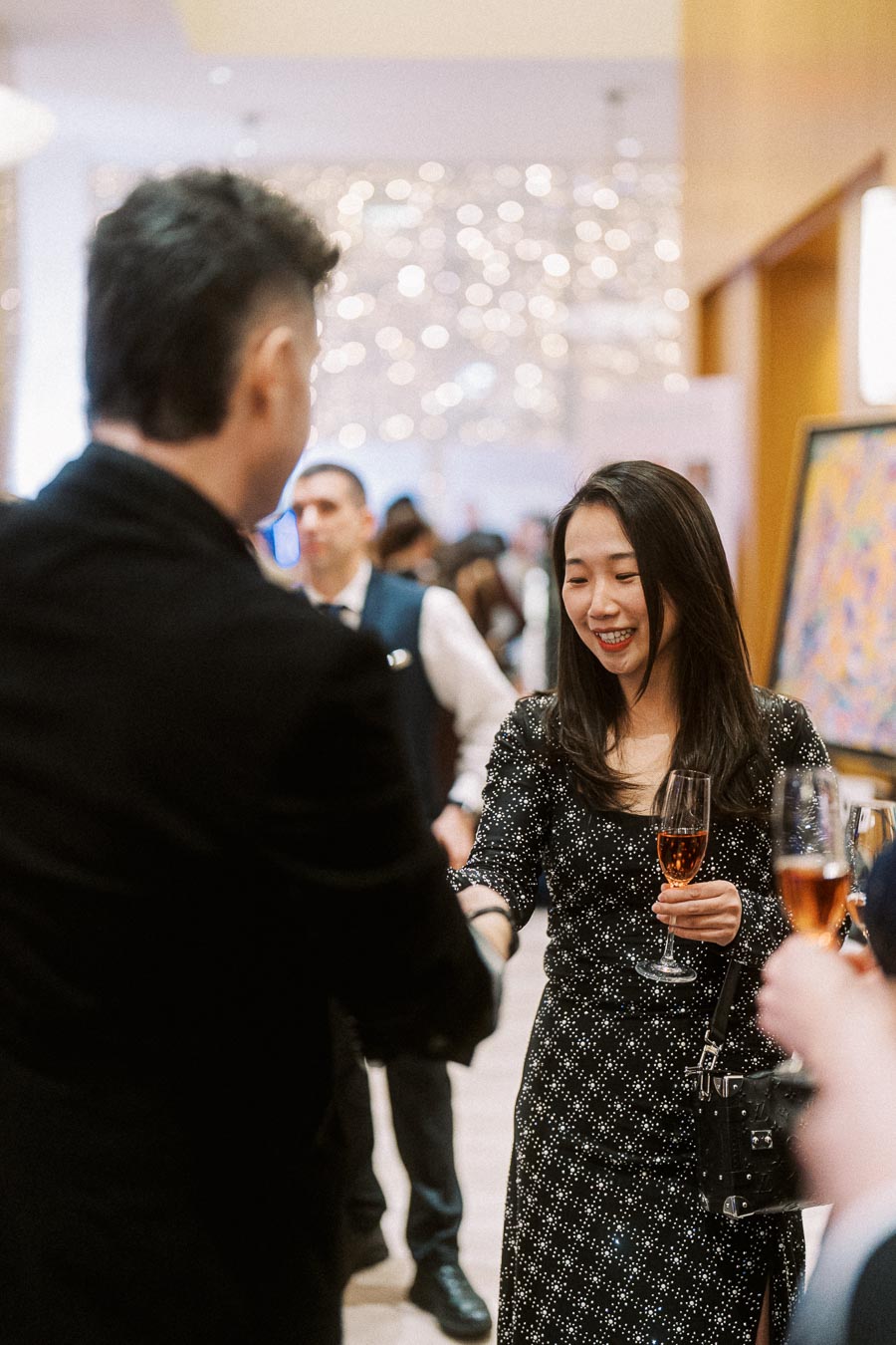 A woman in an elegant black dress holding a glass of champagne, smiling and engaging in conversation at a sophisticated social event. The background features art and a bustling crowd, creating a lively atmosphere.