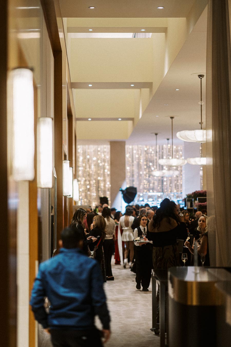A busy, elegant hallway filled with people socializing at a formal event, featuring sophisticated lighting and decor, suggestive of a high-end gathering or reception.