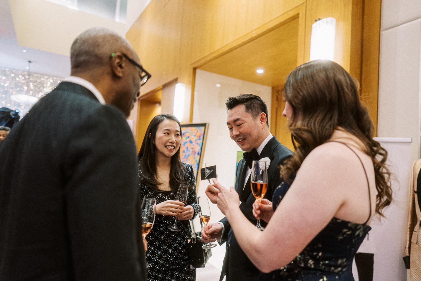Group of people enjoying a social gathering, dressed in formal attire, holding champagne flutes, and engaging in conversation at an elegantly decorated venue.