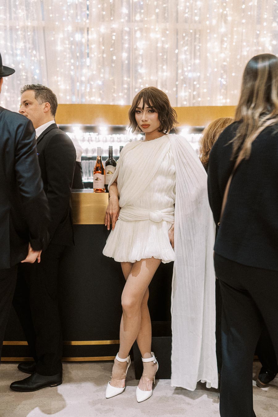 Elegant woman in a stylish white dress poses confidently at a sophisticated cocktail bar, surrounded by people in formal attire, with sparkling lights in the background creating a glamorous atmosphere.