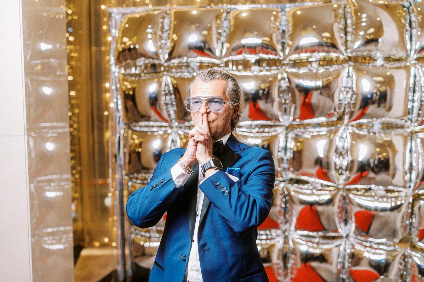 Stylish man in a blue suit and glasses stands pensively in front of a shiny, reflective balloon backdrop at a glamorous event.