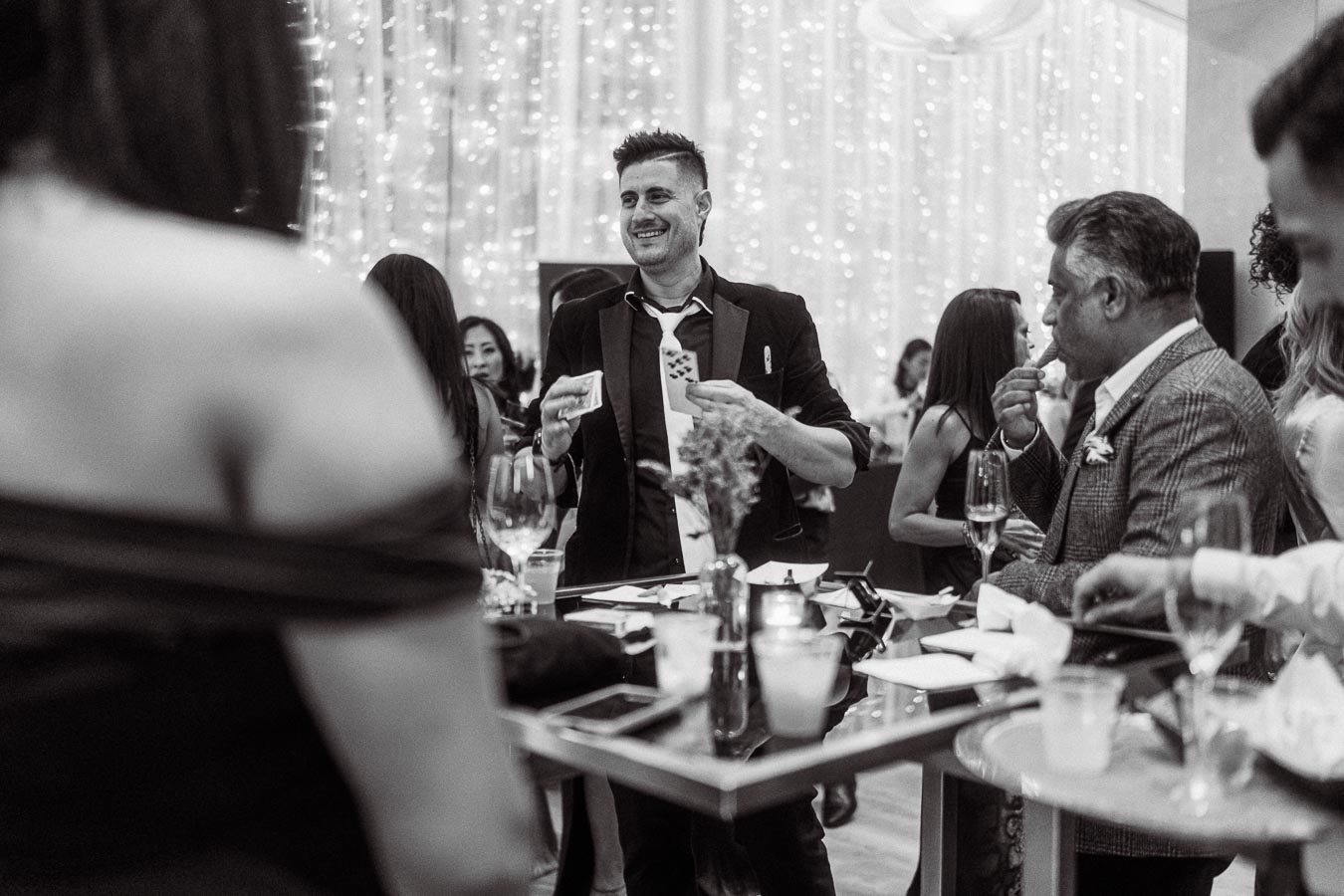 Black and white image of a lively social gathering with elegantly dressed people enjoying themselves around a table at an event, surrounded by sparkling lights.