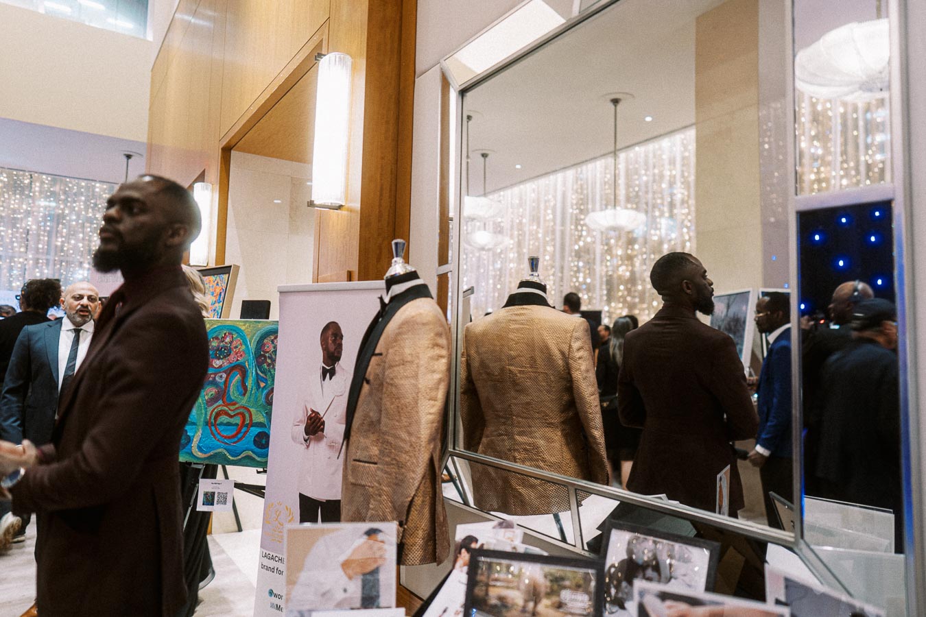 Elegantly dressed individuals at a fashion-themed event, featuring tailored suits on display, a man viewing the exhibit, and a mirrored backdrop enhancing the elegant ambiance.