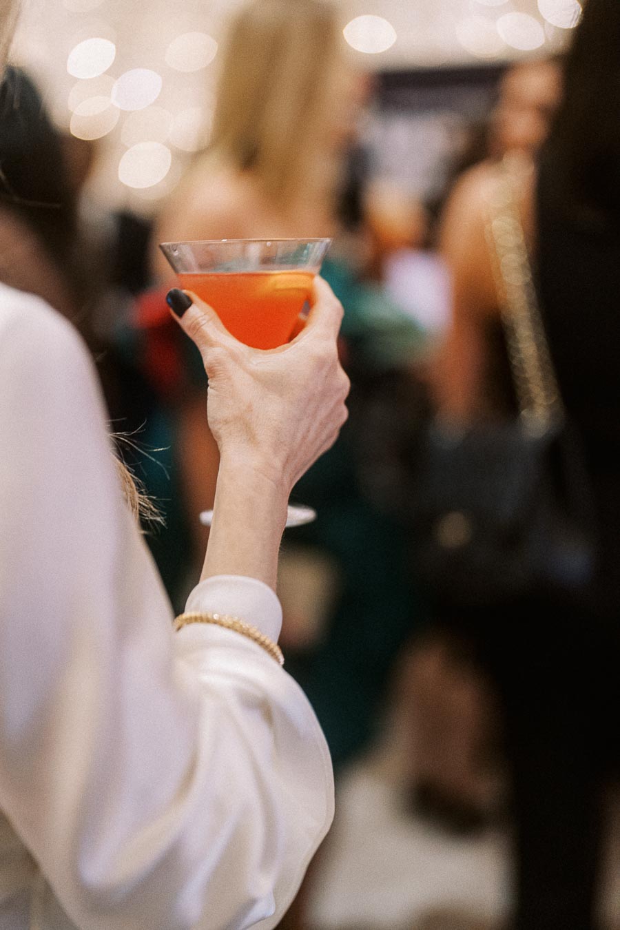 A person holding a red cocktail in a martini glass at a stylish social event, with a blurred background of people mingling.