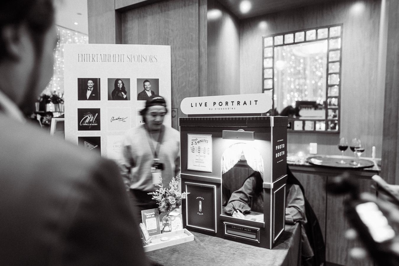 Black and white image of a live portrait photo booth at an event, featuring a display of entertainment sponsors and a person interacting with the booth setup.