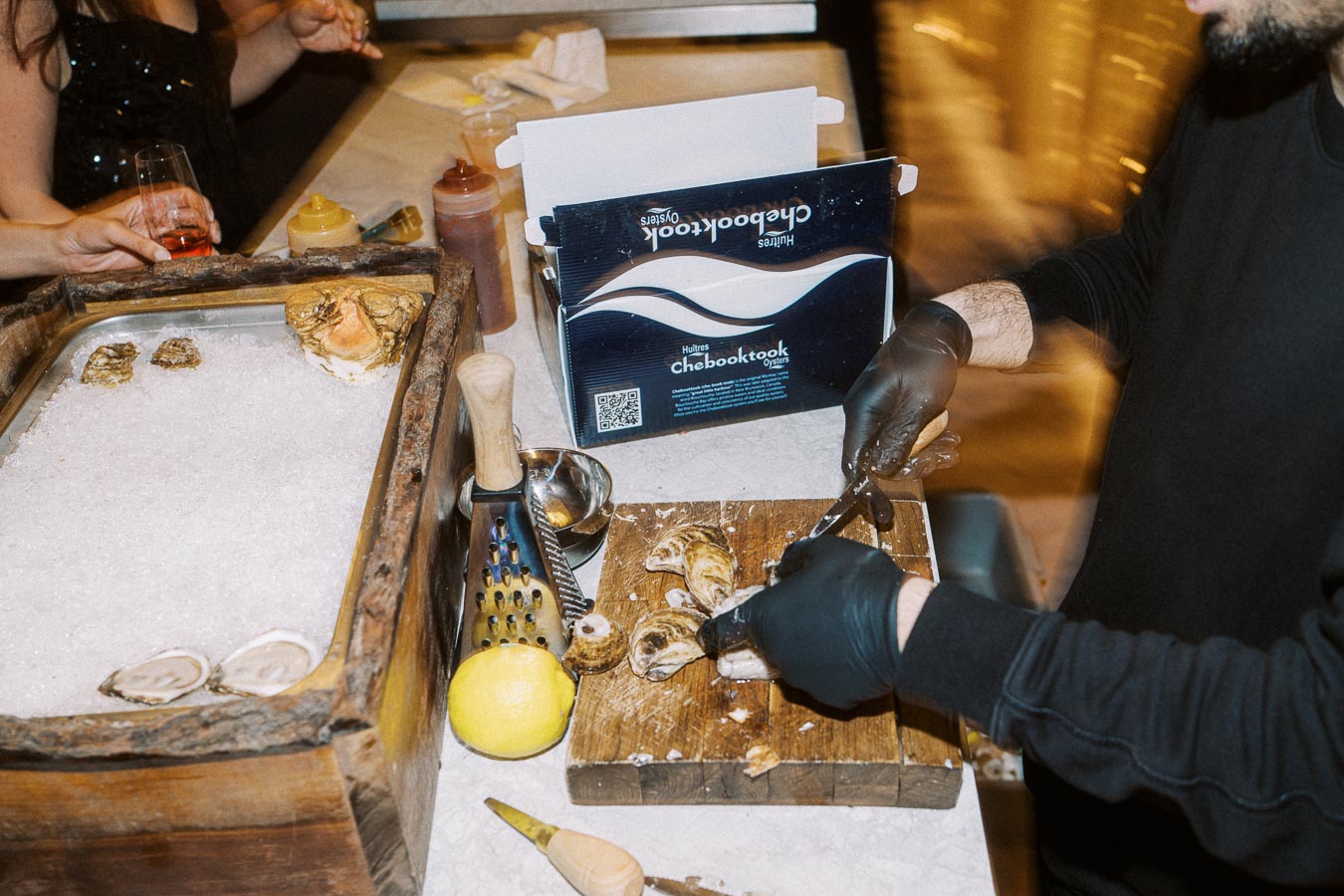 Oyster shucking demonstration at a seafood event. A person wearing gloves uses a knife to open oysters on a wooden board, with a tray of ice and opened oysters nearby. A box labeled Chebooktook Oysters and various condiments are visible in the background.