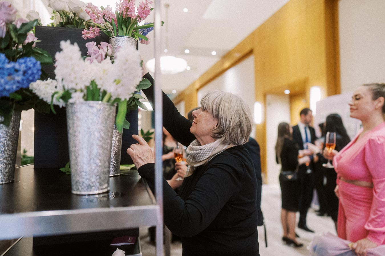 A woman arranging flowers in silver vases at an elegant indoor event with attendees in formal attire in the background.