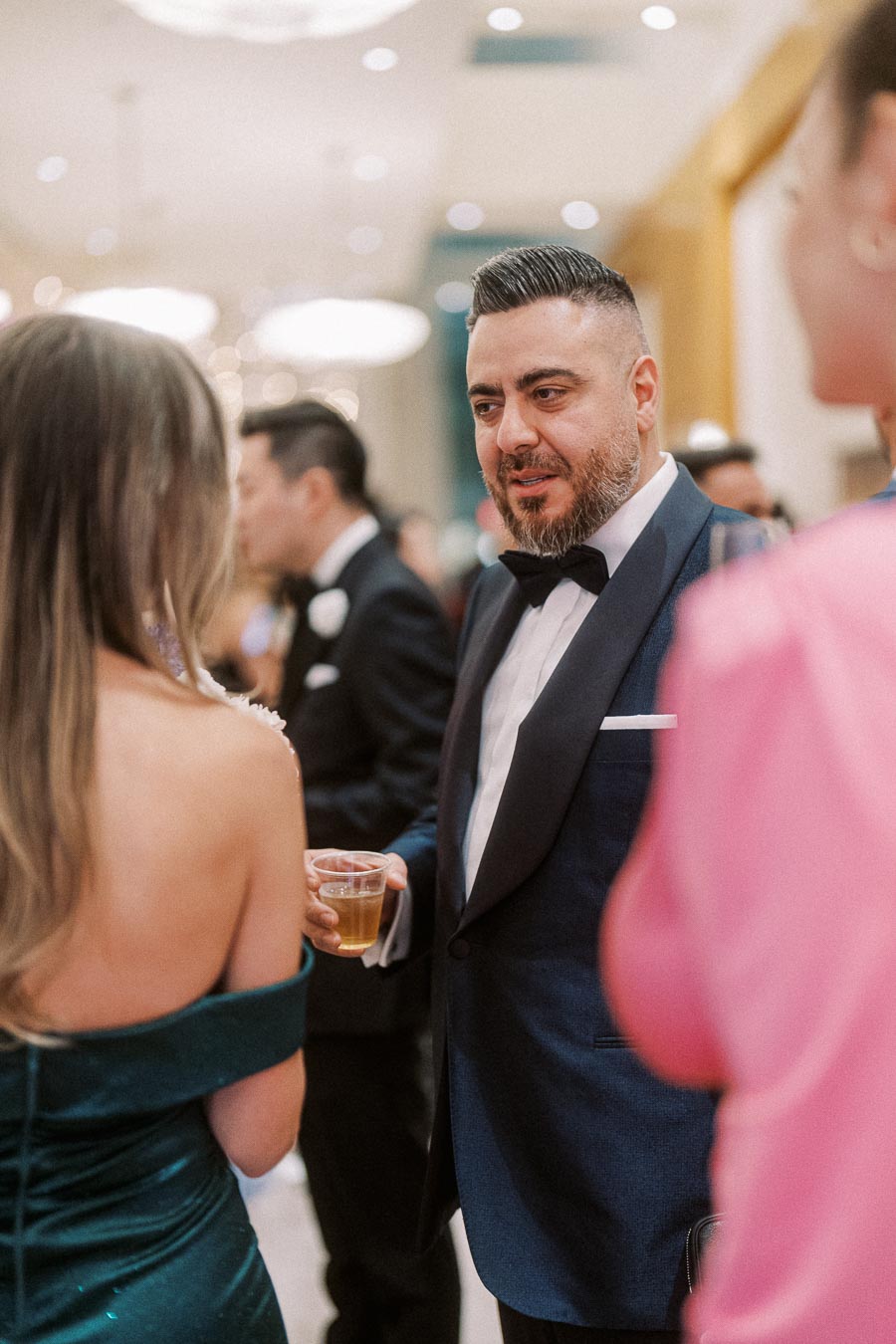 A man in a dark suit and bow tie socializes at an elegant event, holding a drink, surrounded by other formally dressed attendees in a warmly lit venue.