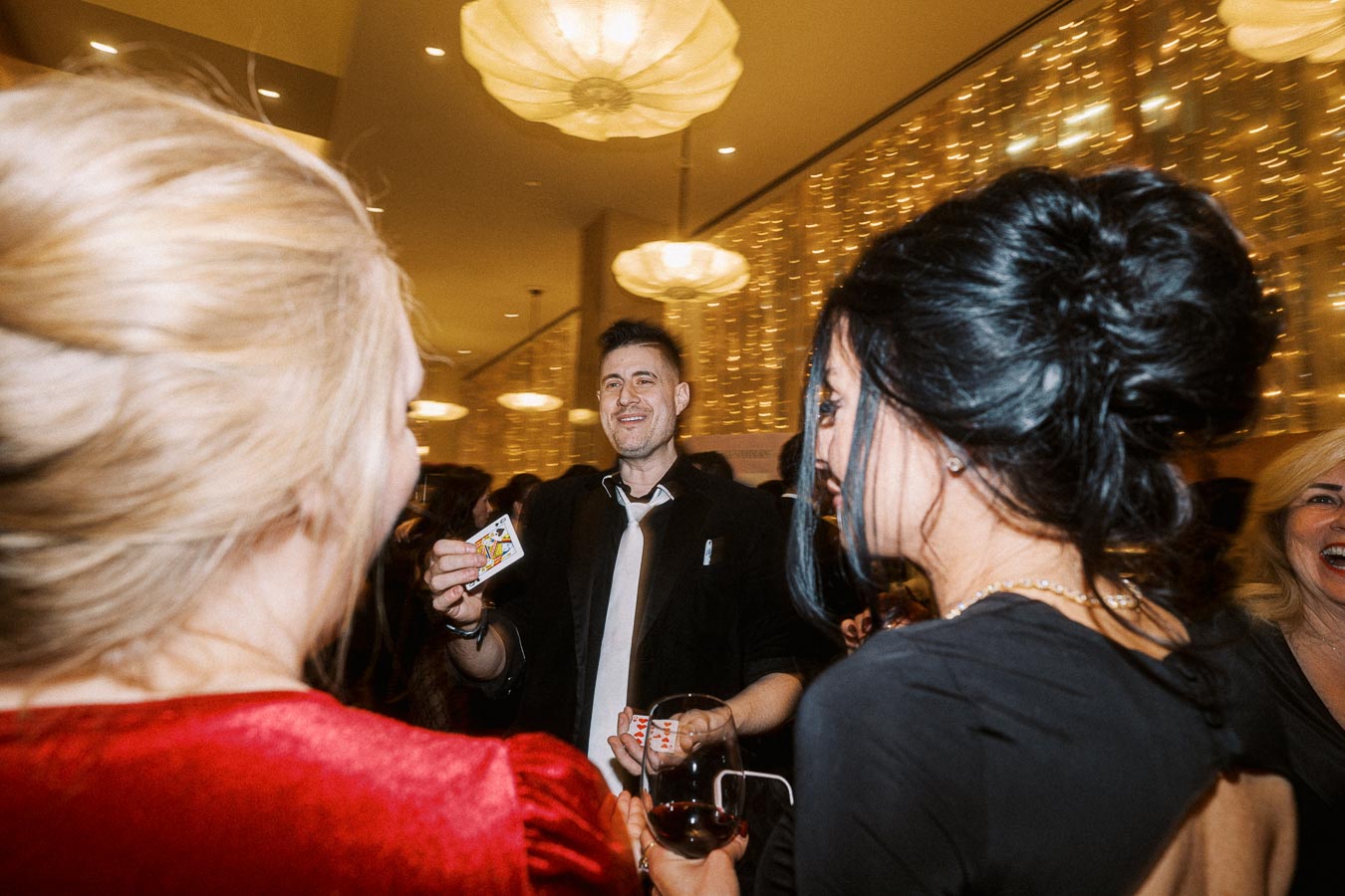 Magician performing a card trick at a lively party, surrounded by an engaged audience in a warmly lit venue with festive decorations.