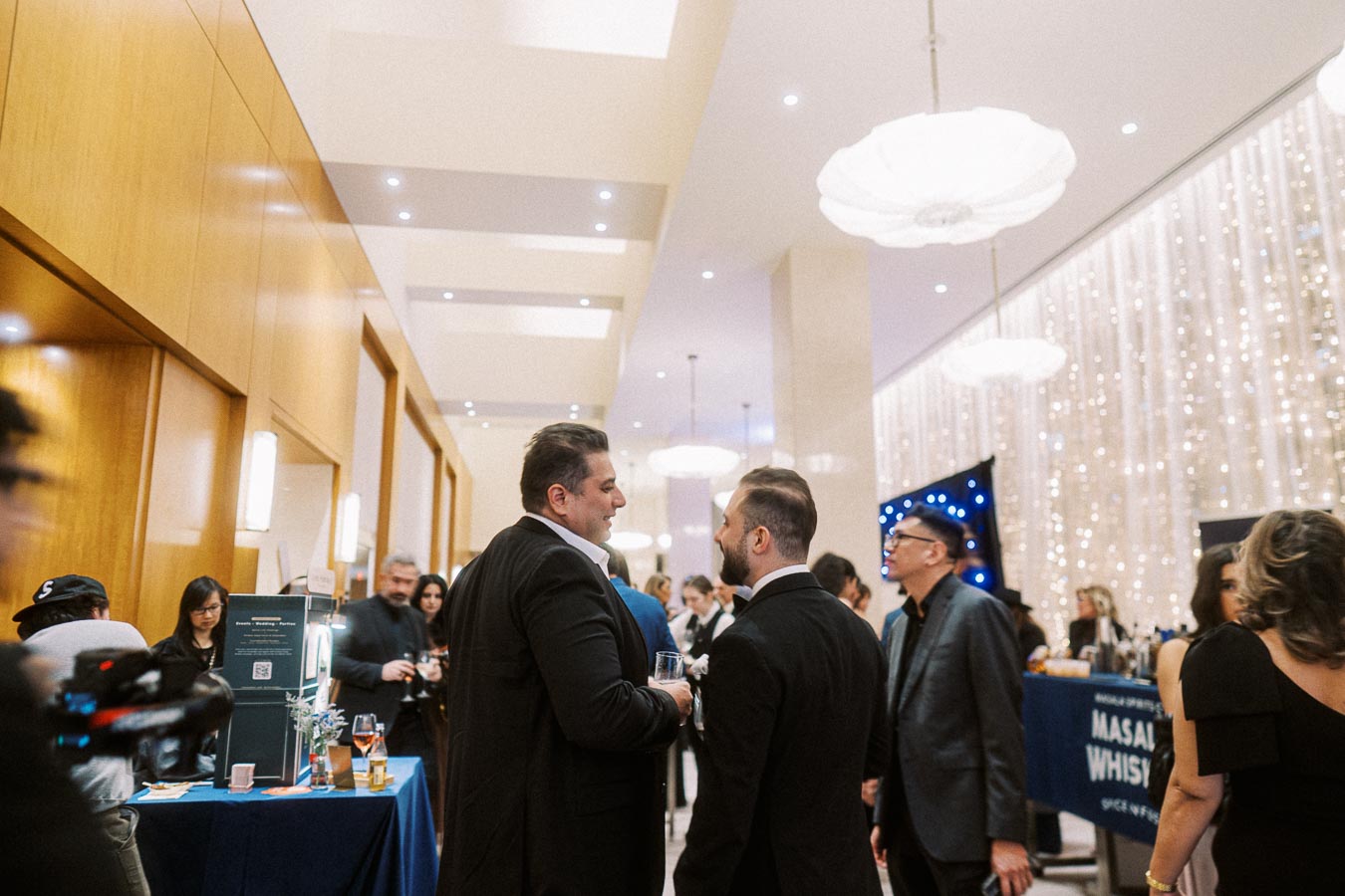 Business professionals networking at a formal indoor event, featuring elegant decor and bright lighting. Tables with promotional materials and attendees engaged in conversation are visible.