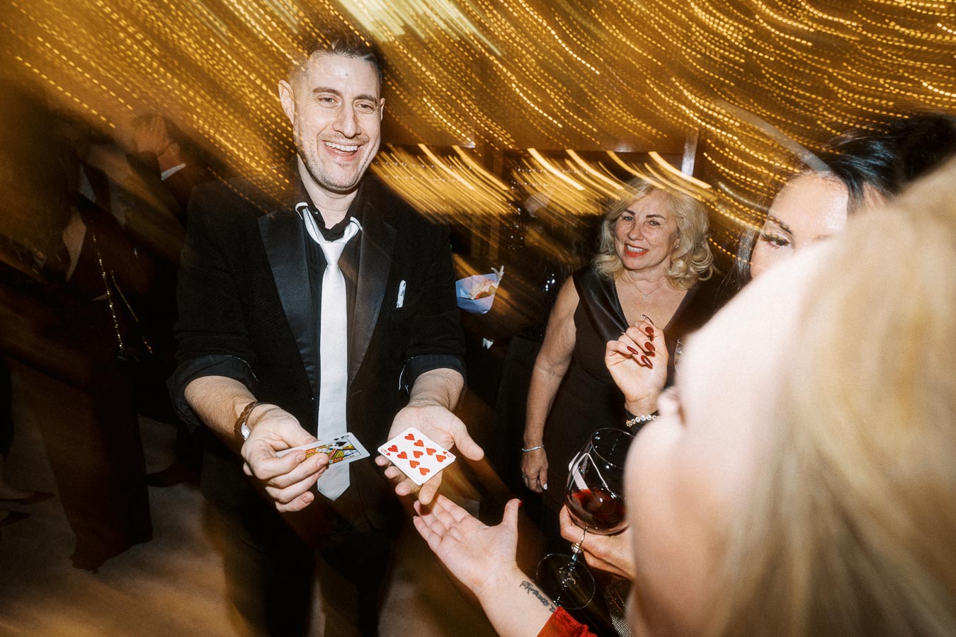 A man in a suit performing a card trick at a lively celebration, surrounded by smiling guests with glasses of wine, against a backdrop of warm, blurred lights.