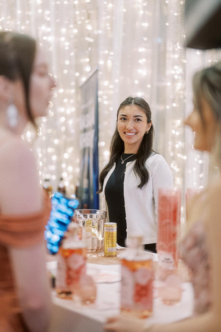 A woman smiling behind a bar setup at a festive event with twinkling lights, engaging with attendees.