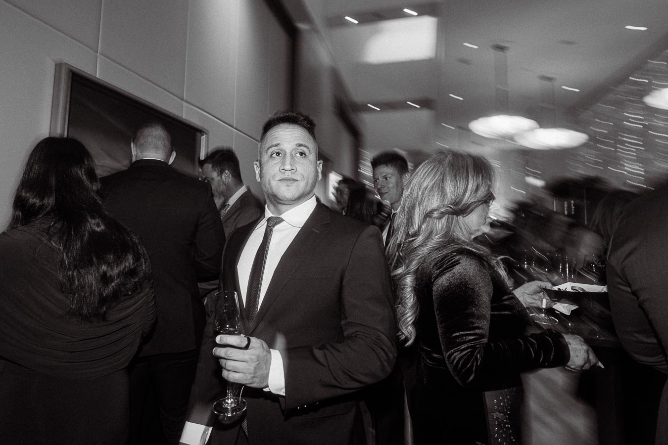 Black and white image of a man in a suit holding a drink at a formal event, surrounded by elegantly dressed guests in a modern, illuminated venue.