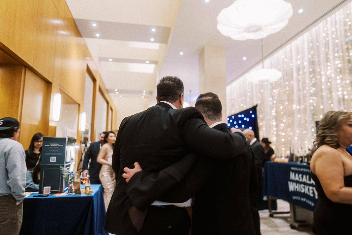 Two people in formal attire embracing at an elegant indoor event, with lit decorations and various attendees mingling in the background.