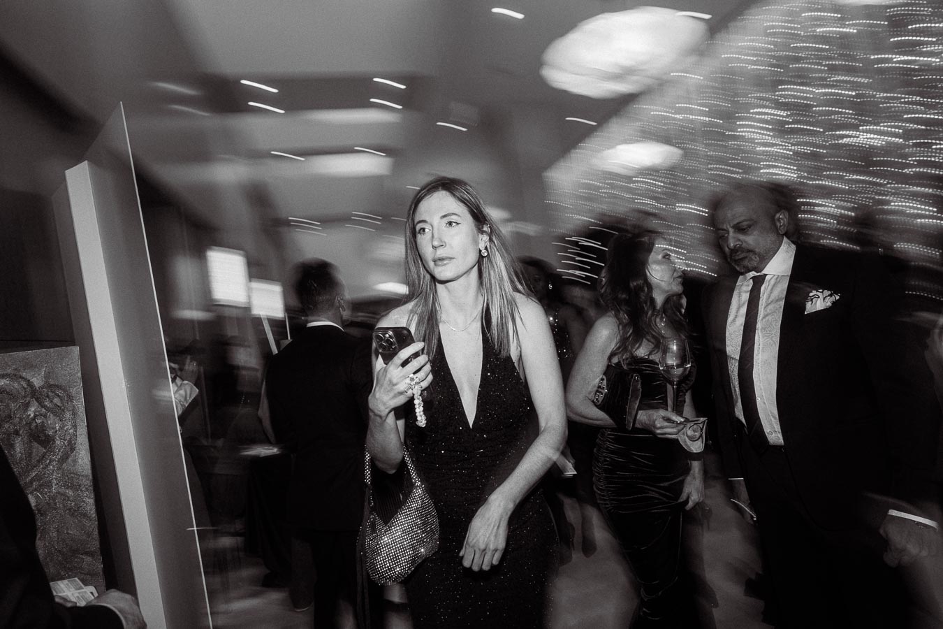 Black and white photo of a stylish event, capturing a woman in a black evening dress holding a smartphone, as she walks through a glamorous, blurred setting with other elegantly dressed guests in the background.