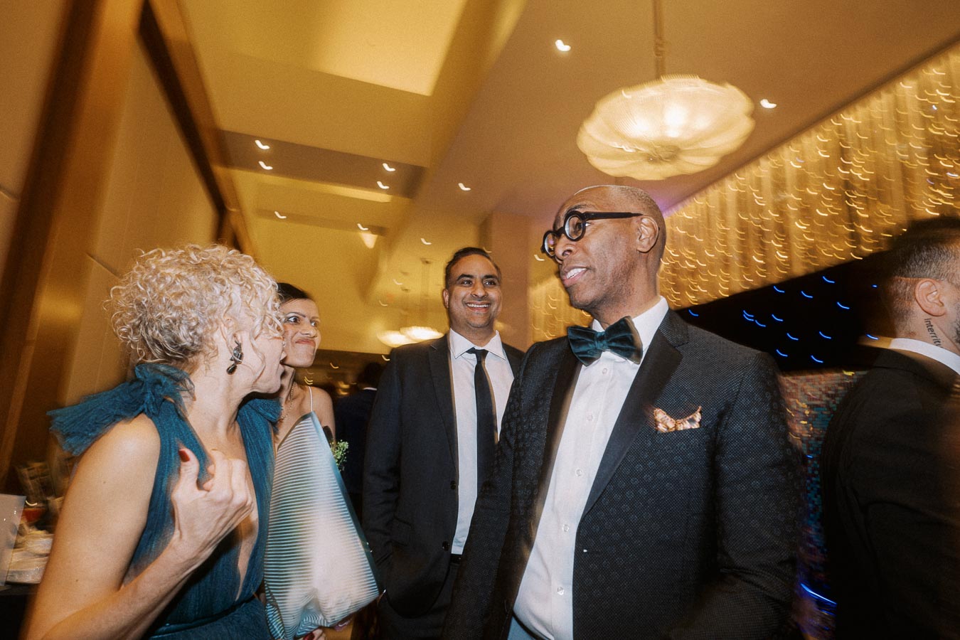 Group of elegantly dressed people having a lively conversation at a formal event; indoor setting with decorative lighting in the background.