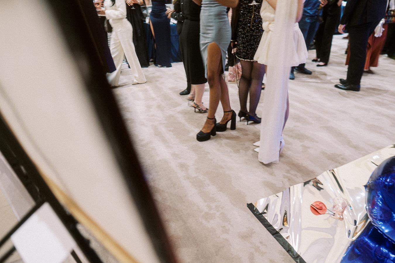 Elegant gathering with attendees in formal attire and high heels, standing on a plush carpet at a stylish event, with reflections from a metallic surface.