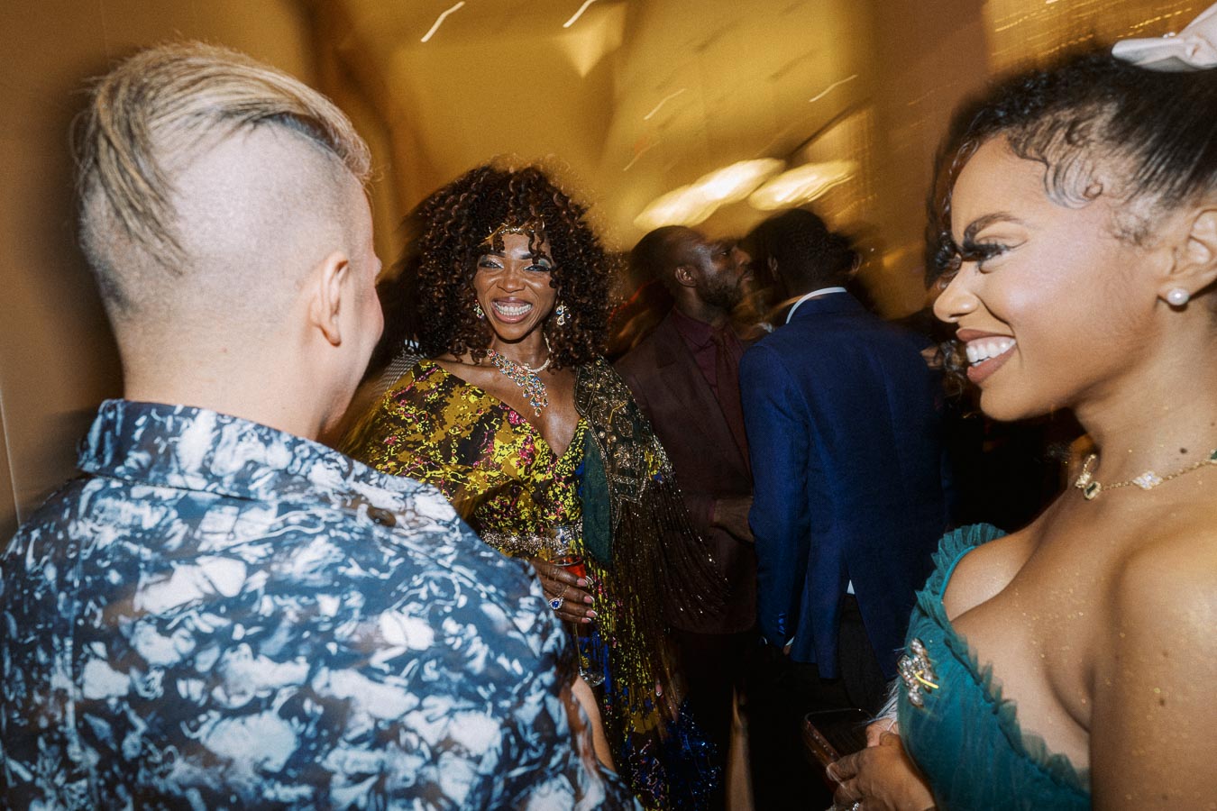 A lively group of elegantly dressed people smiling and conversing at a social event, illuminated by warm lighting, creating a vibrant and joyful atmosphere.