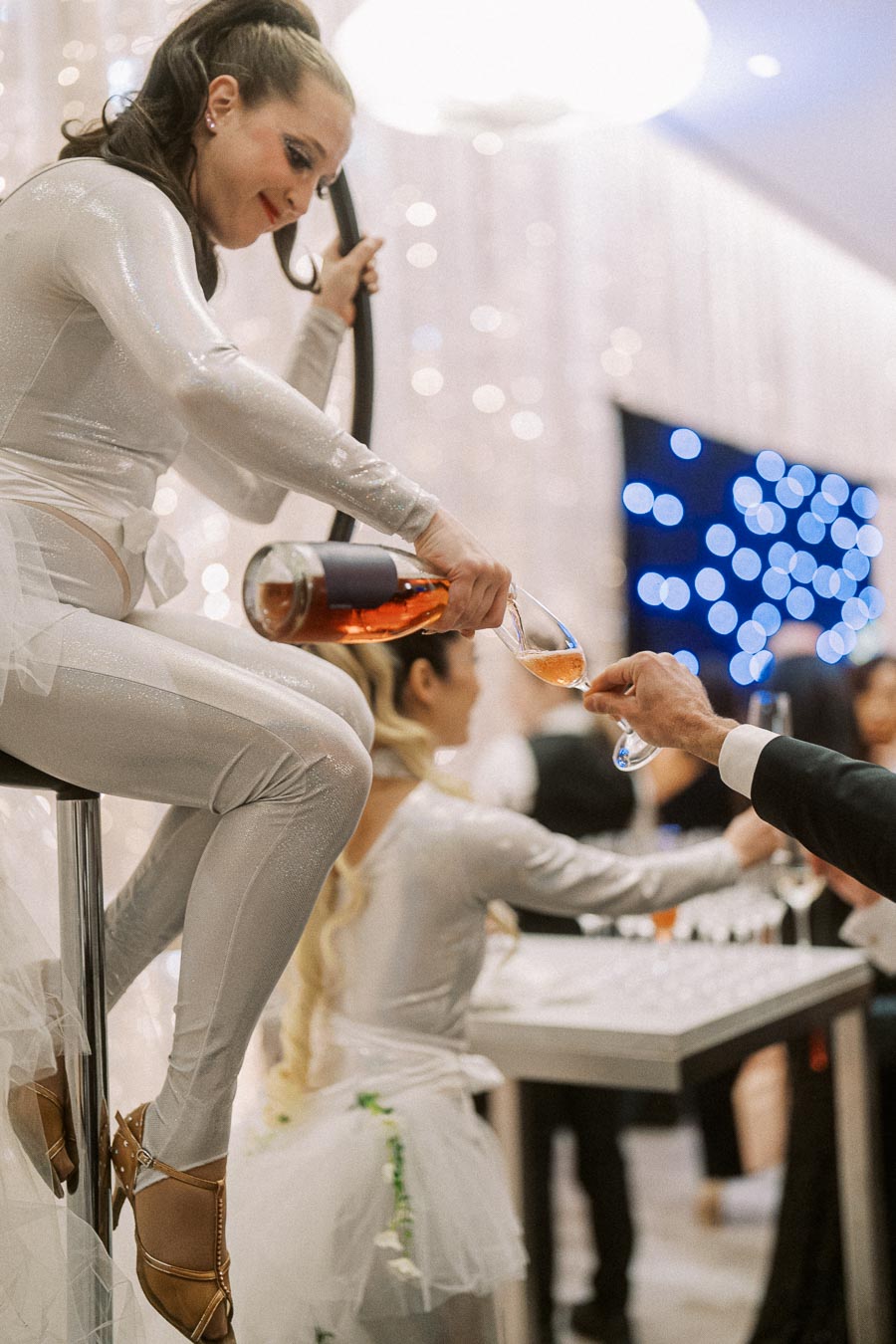 Aerial performer in a silver costume pouring wine at an elegant event, with sparkling lights and attendees in the background.