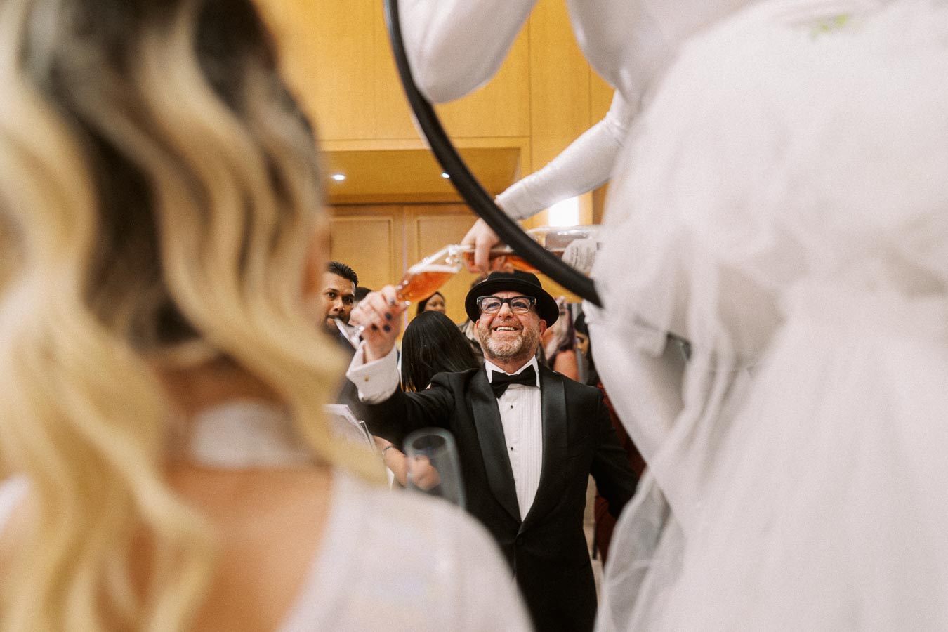A man in a tuxedo and hat toasting with a champagne flute during a festive celebration, with a blurred woman in the foreground and a performer on stilts in the background.