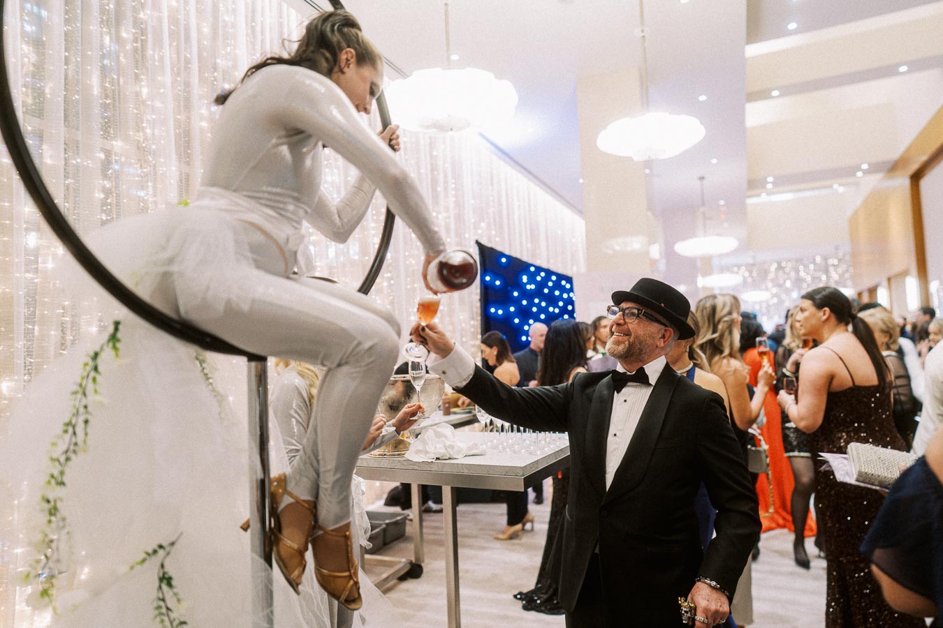 Aerial performer pouring wine into a glass held by a well-dressed guest at a glamorous, upscale event with elegant lighting and a festive atmosphere.