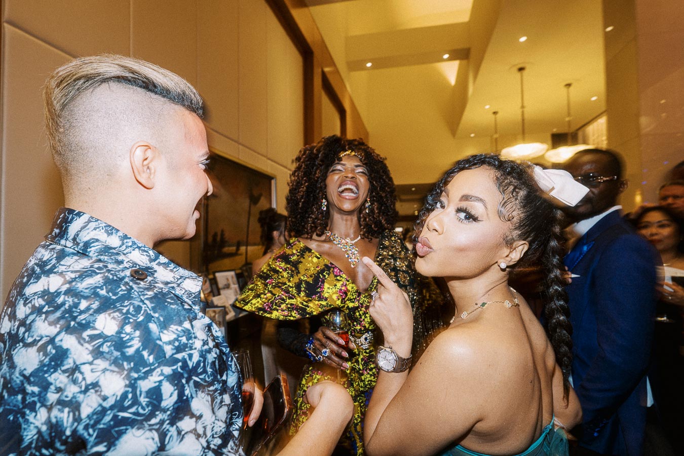 A lively group of elegantly dressed individuals enjoying a lively party, featuring a woman in a vibrant patterned dress laughing, a woman with braided hair making a playful pout, and a person with a stylish haircut in a patterned shirt, all gathered in a warmly lit, sophisticated setting.