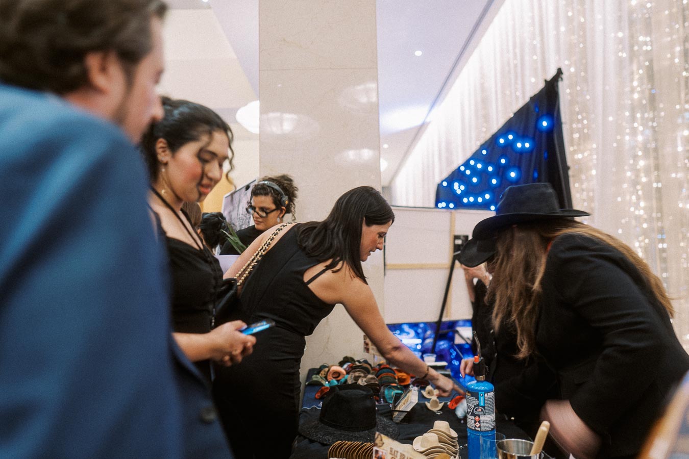 Group of people browsing handmade accessories at a market stall, with festive lights in the background.