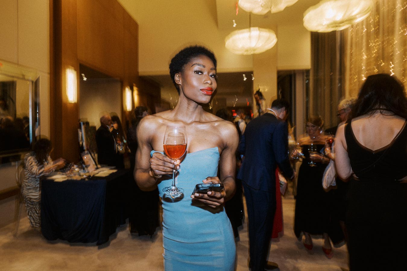 Female attendee in a stylish blue dress enjoying a drink at a sophisticated evening event, surrounded by elegantly dressed guests in a warmly lit venue.