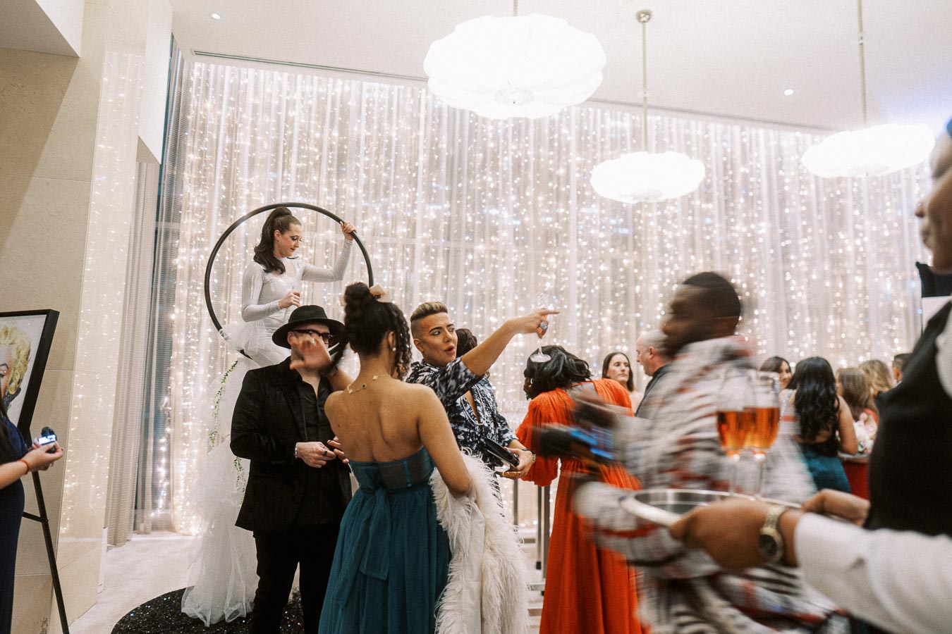 A lively party scene with elegantly dressed guests socializing in a room decorated with twinkling lights. A performer in white gracefully balances on a hoop, adding to the festive atmosphere. A waiter carrying drinks moves through the crowd. The setting suggests a glamorous and upscale event.