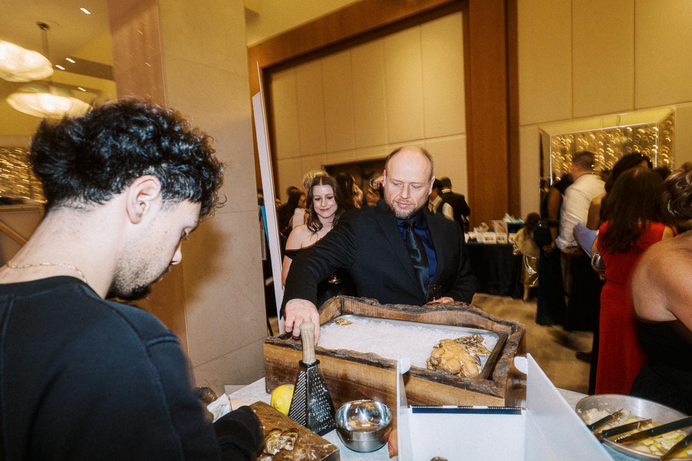 A man in a suit interacts with an oyster bar at a formal event, surrounded by elegantly dressed guests in a well-lit banquet hall.