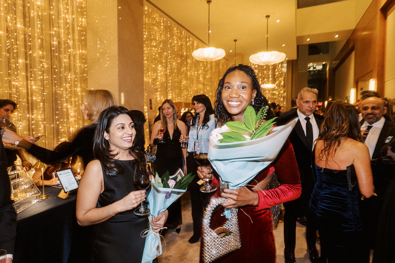 Group of elegantly dressed individuals enjoying a sophisticated cocktail party, featuring warm ambient lighting, floral bouquets, and joyous expressions.
