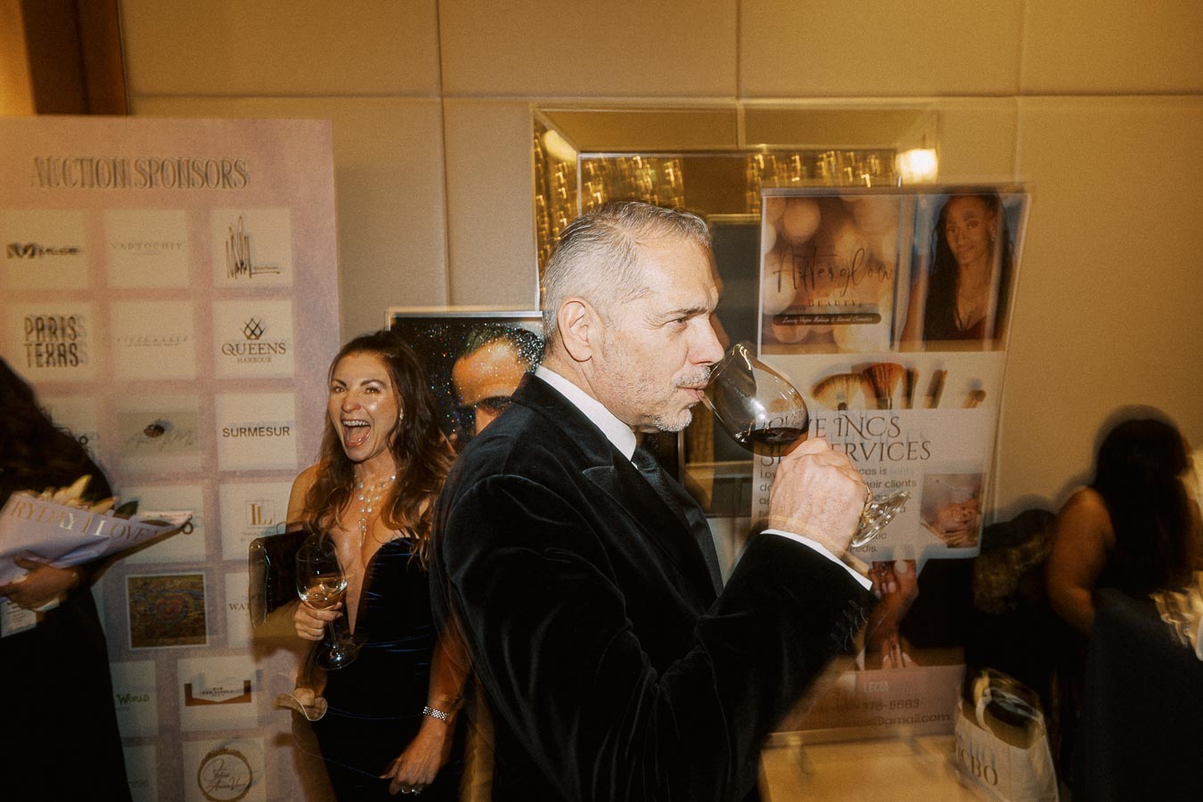Elegant event scene with a man in a black suit savoring a glass of wine, surrounded by smiling guests and promotional sponsor displays in the background.