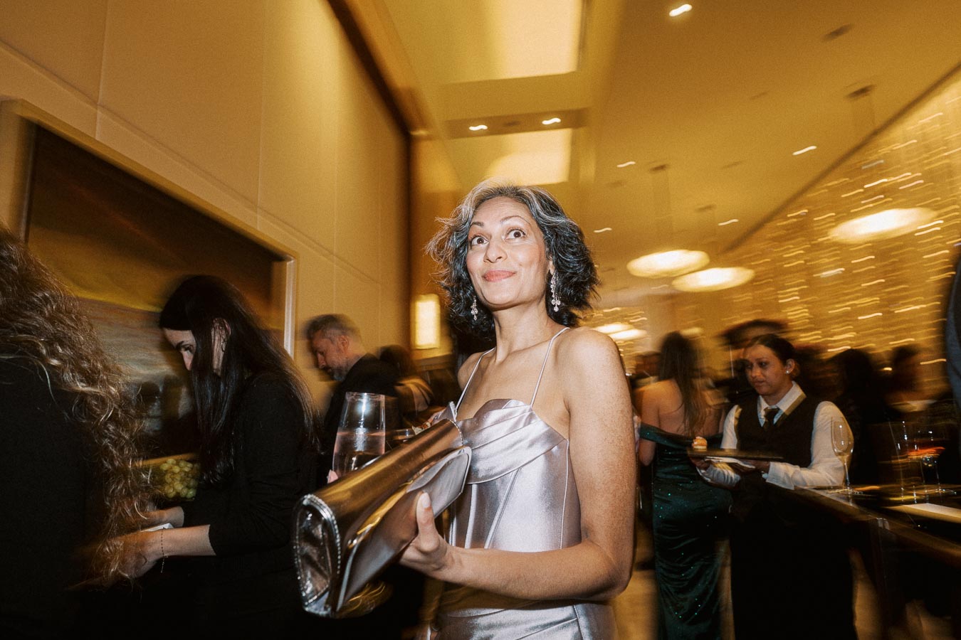 Elegant woman in silver dress holding a clutch and glass, smiling at a formal event with a blurred crowd and warm lighting in the background.