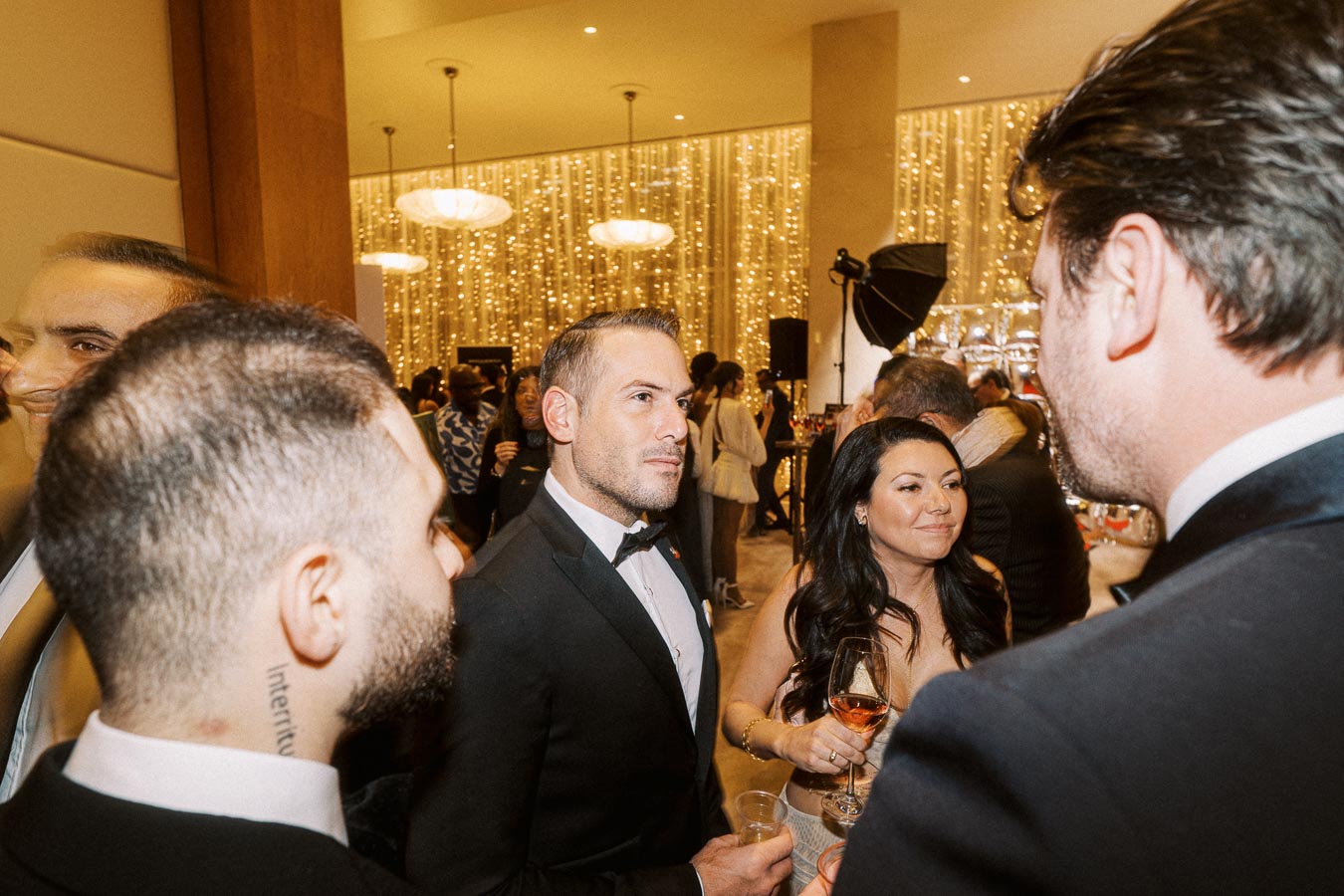 A group of elegantly dressed individuals socializing at a formal event with sparkling lights in the background, featuring men in tuxedos and a woman holding a glass of wine, creating a sophisticated and lively atmosphere.