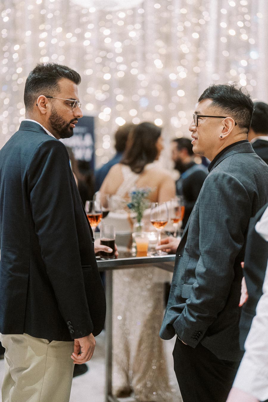 Two men in suits engaging in conversation at an elegant social gathering, with a bokeh backdrop and drinks on a table in the foreground.