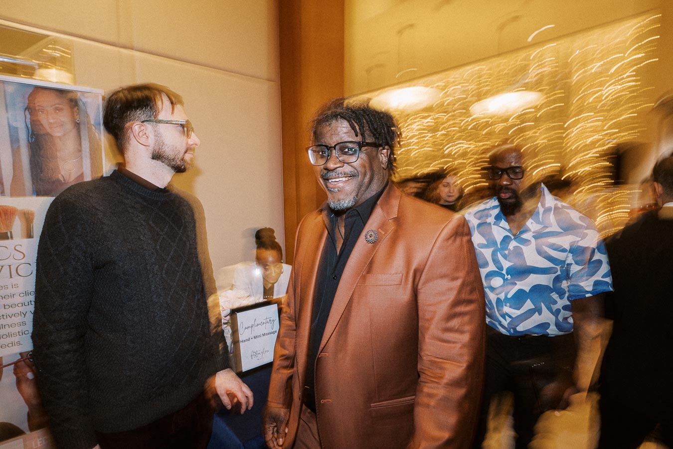 A group of people enjoying a lively event. A man in a stylish orange suit is smiling, surrounded by a buzzing atmosphere with blurred lights. A man in a blue patterned shirt and another in a dark sweater are nearby, encapsulating a sense of camaraderie and celebration.
