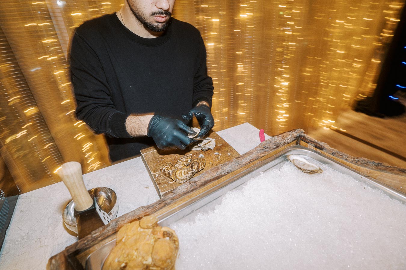 Man shucking oysters on a wooden board at an upscale event with sparkling golden lights in the background.