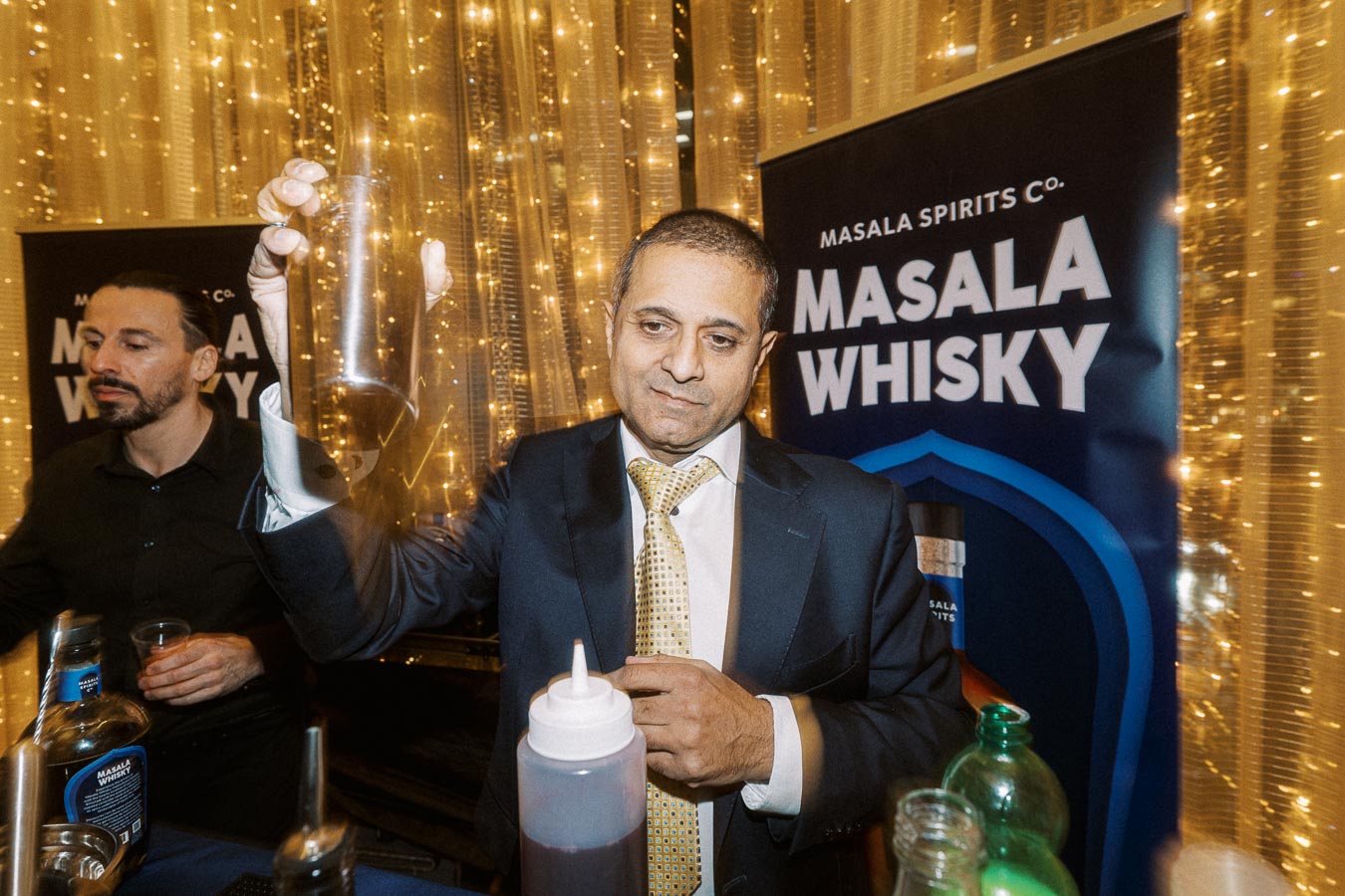 A well-dressed man holding a cocktail shaker behind a bar with a Masala Whisky banner in the background, featuring festive lighting.