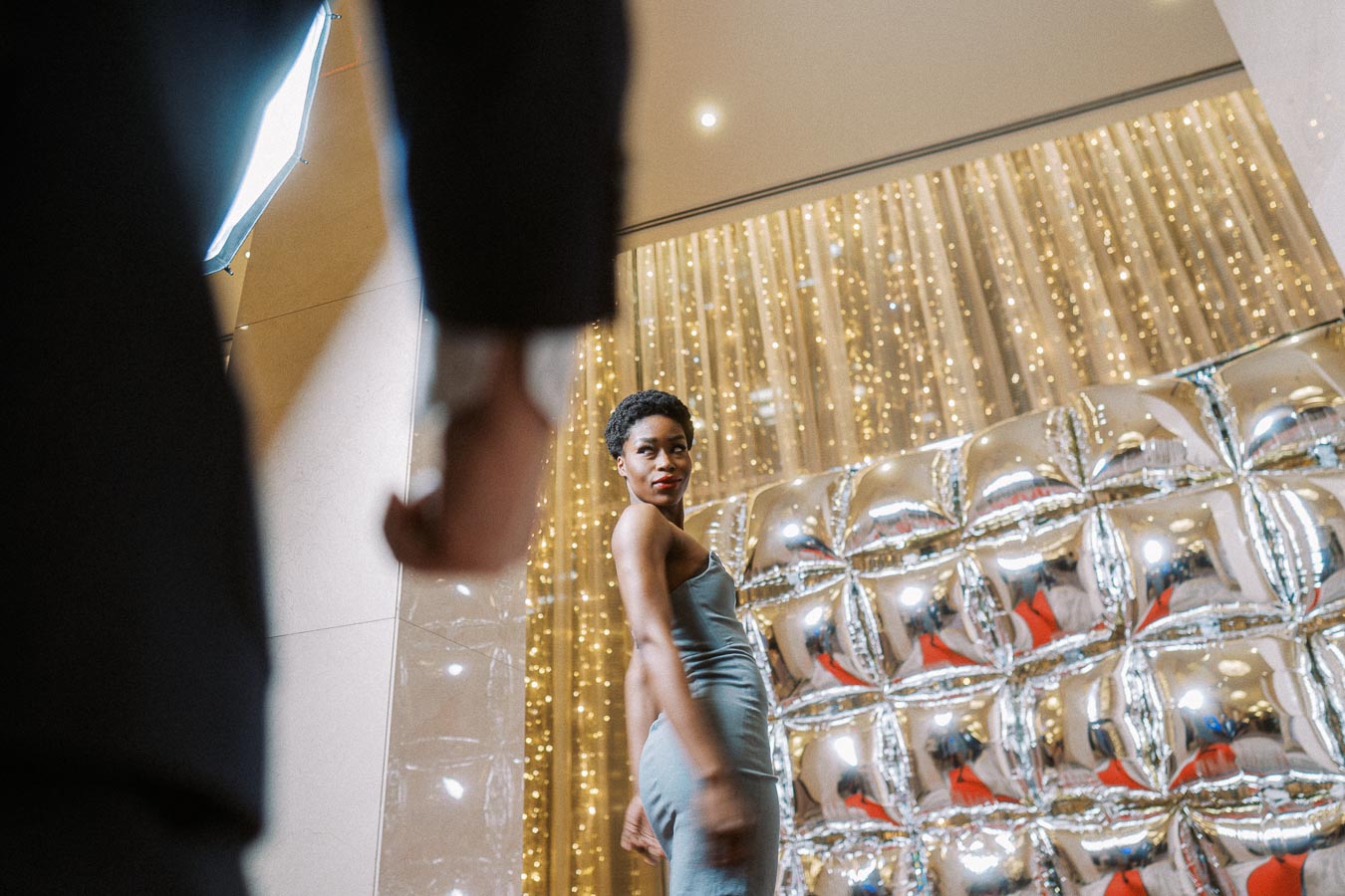 Glamorous woman in a sleek dress posing elegantly in front of a wall adorned with shiny, metallic balloons and twinkling lights, capturing a sophisticated ambiance.