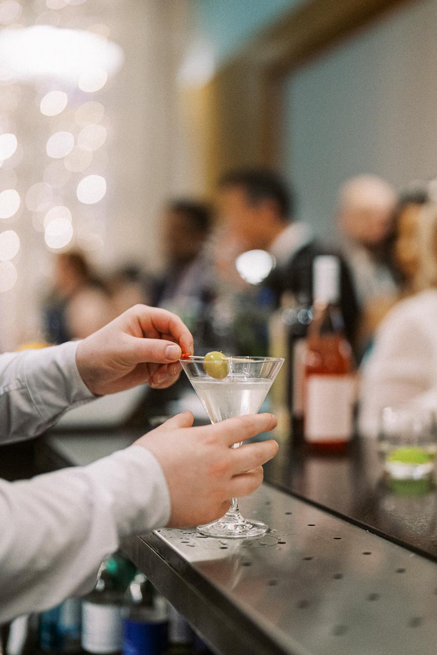 Person holding a martini glass with an olive garnish in a social gathering at a bar, with blurred people in the background.