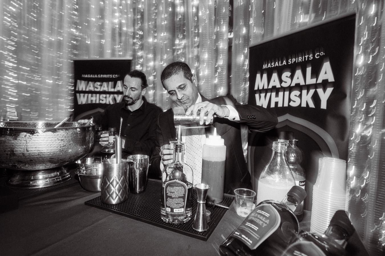 Bartender crafting cocktails with Masala Whisky in an elegant setting, featuring shimmering lights and promotional banners for Masala Spirits Co.