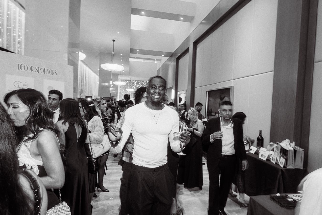 A lively black and white photo captures a smiling man enjoying a social event, holding a drink in each hand. He is surrounded by a well-dressed crowd engaging in conversation, set in a spacious, modern venue with high ceilings and elegant lighting.
