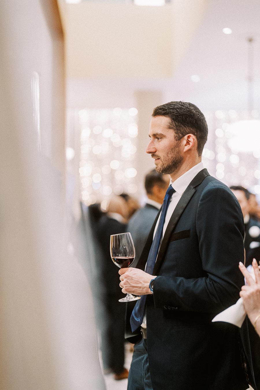 Man in a suit holding a wine glass at an elegant social event, surrounded by blurred ambient lights and other guests.