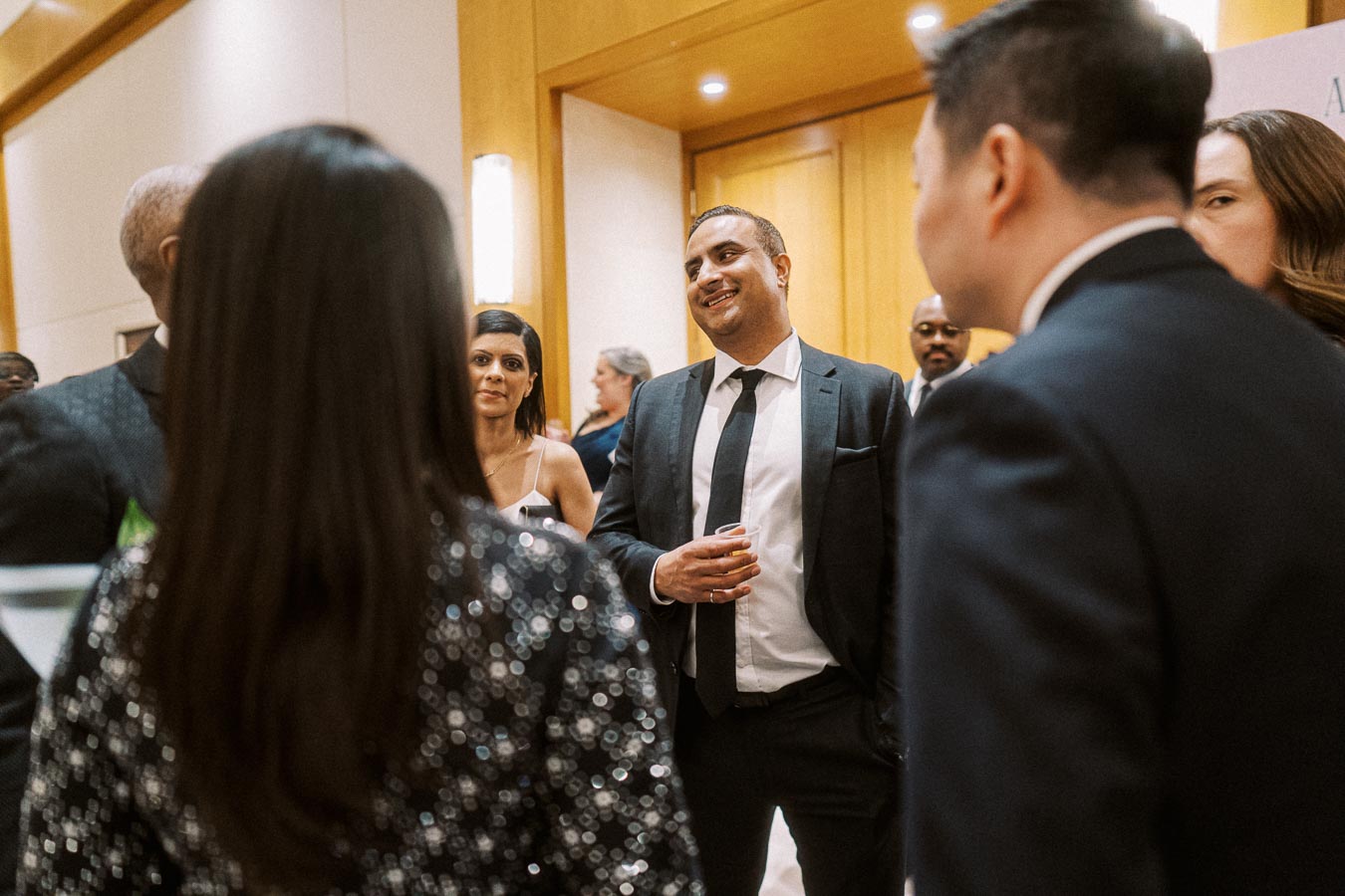 Group of people networking at a formal event, dressed in business attire, engaged in conversation with a warm and lively atmosphere.
