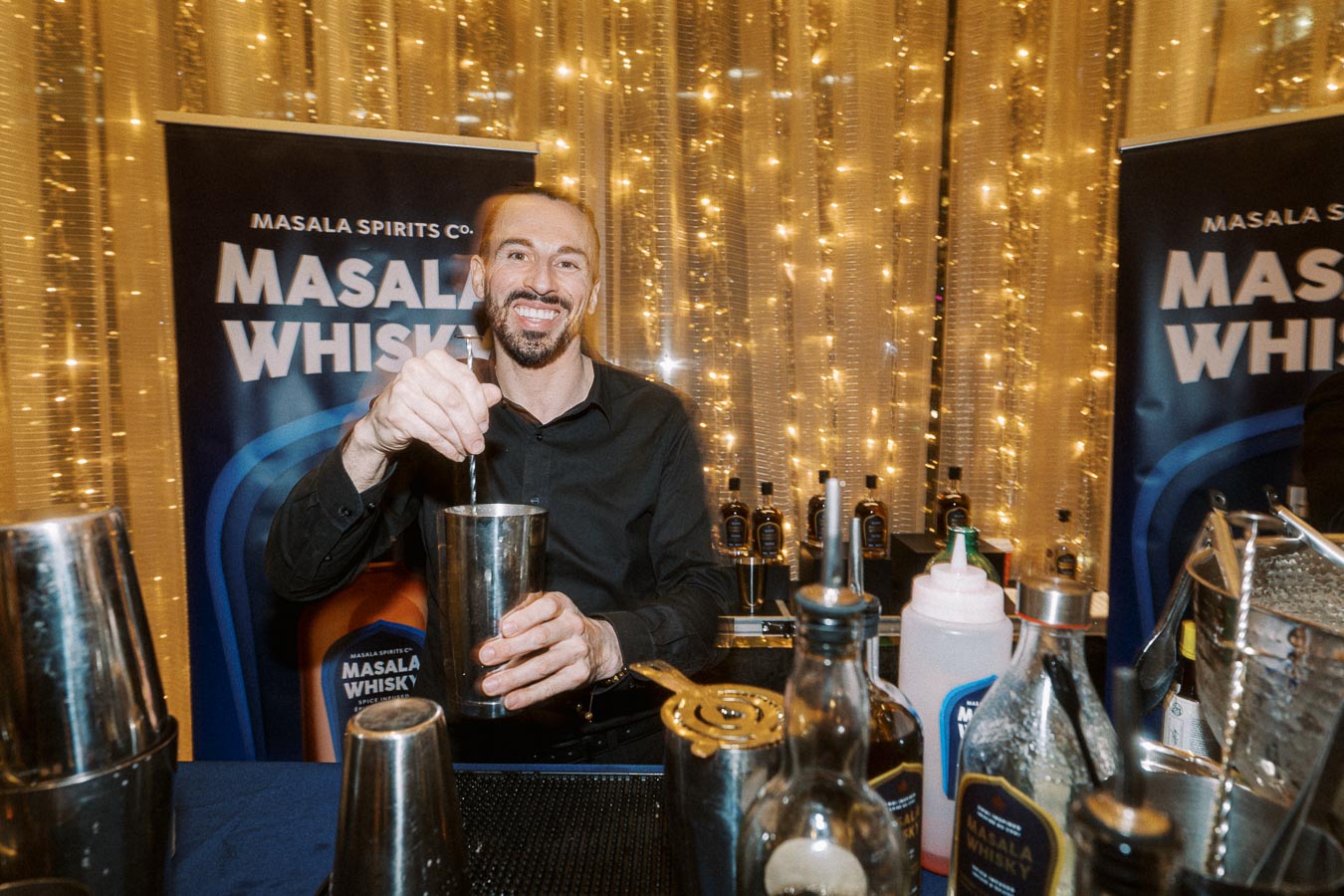 Bartender preparing a cocktail at a Masala Spirits Co. event, surrounded by bottles of Masala Whisky, against a backdrop of warm, decorative lights.
