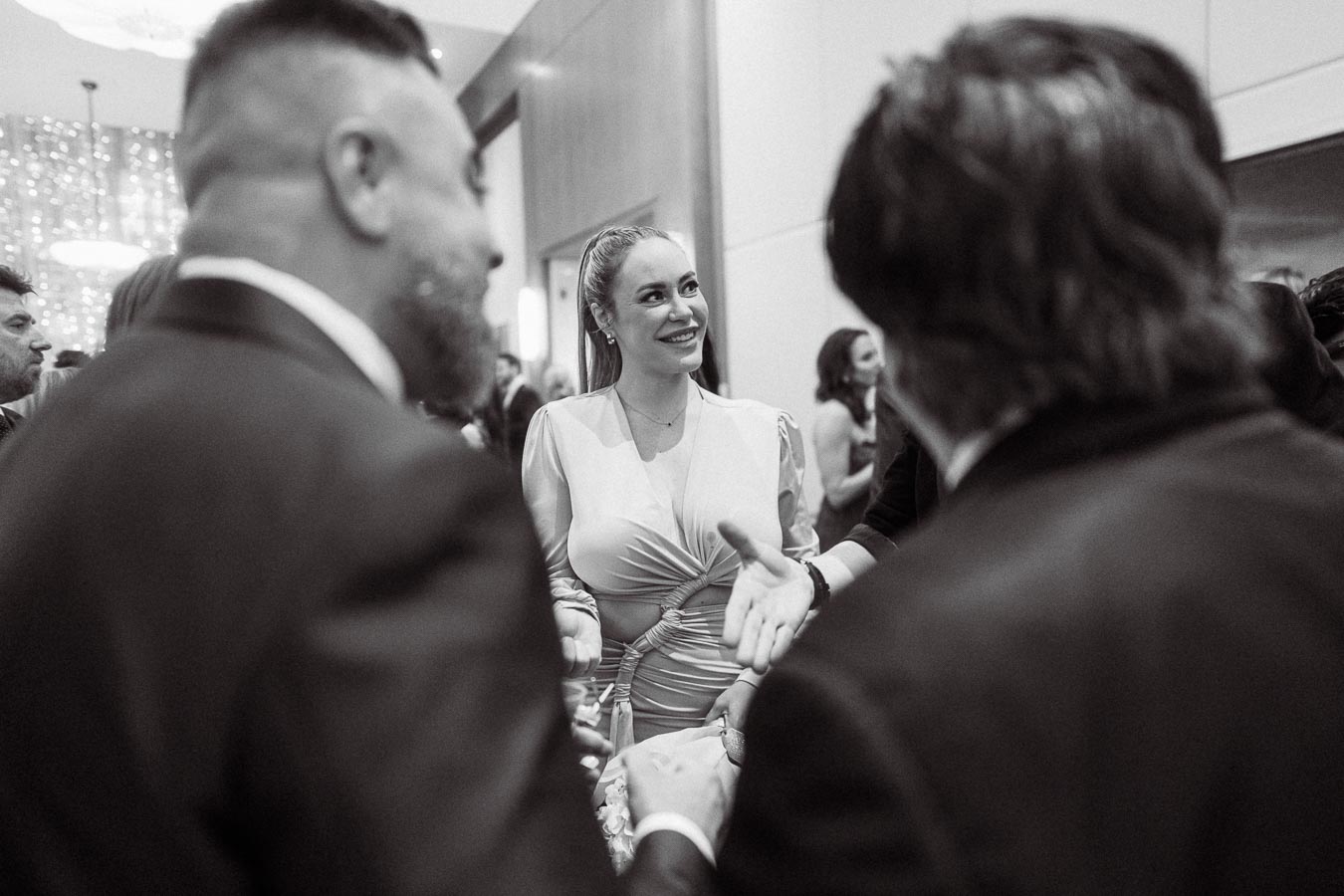 Black and white photo of a smiling woman at a social event, engaged in conversation with two men. The setting appears to be a formal gathering, with elegantly dressed attendees and a softly lit ambiance.
